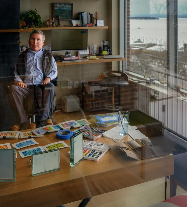 Ned Church at his desk with postcards and paints, seen through the studio window